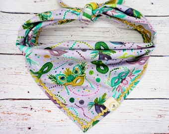 May include: A lavender dog bandana with a repeating pattern of Mardi Gras masks in green, purple, and gold. The bandana is trimmed with gold trim and tied at the top. A small tag is sewn on the bottom corner.