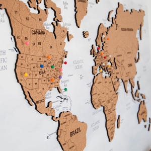 Peut inclure: Une carte du monde en liège avec les contours des pays et des punaises colorées marquant les lieux. La carte comprend les États-Unis, le Canada, le Mexique, le Brésil et le Groenland. La carte est montée sur un fond blanc.