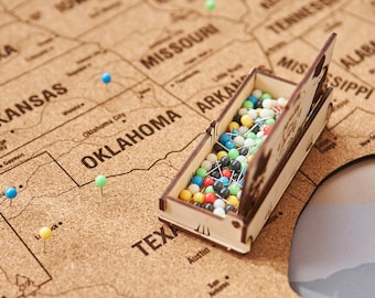 May include: A corkboard map of the United States with push pins in various states. A wooden box filled with colorful push pins is open and sitting on top of the map.