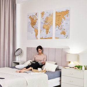 May include: A woman is sitting on a bed with a grey blanket and white pillows, reading a book. She has a tray with a cup and a plate on her lap. There are three world maps on the wall behind her, each featuring a different continent. The maps are made of wood and have a light brown colour.