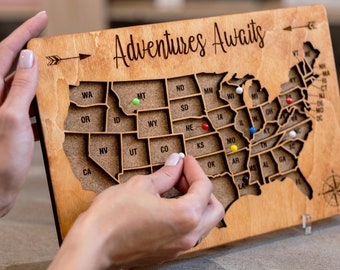 Könnte beinhalten: Eine Holzkarte der Vereinigten Staaten mit den Worten "Adventures Awaits" oben eingraviert. Die Karte hat Aussparungen für jeden Bundesstaat und ist so konzipiert, dass sie mit Stecknadeln verwendet werden kann, um besuchte Orte zu markieren. Eine Hand platziert eine weiße Stecknadel auf dem Bundesstaat Colorado. Eine kleine Holzkiste mit dem Wort "Pins" auf dem Deckel befindet sich in der unteren rechten Ecke des Bildes. Die Kiste enthält eine Vielzahl von farbigen Stecknadeln.