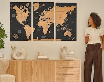 May include: Three black framed world maps with gold foil accents hang on a white wall above a light wood dresser. The maps are divided into three sections: North America, Europe, and Asia. A woman stands to the right of the maps, wearing a white blouse and brown pants.