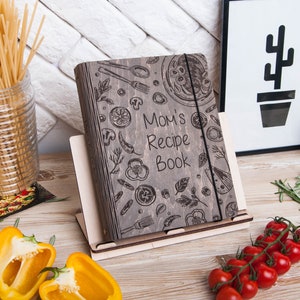 May include: A gray wooden recipe book with a black elastic band closure. The cover features a hand-drawn design of various food items, including vegetables, herbs, and a pizza. The text "Mom's Recipe Book" is engraved on the cover.