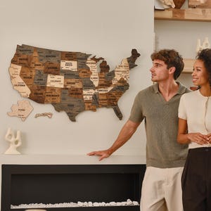 Könnte beinhalten: Eine Holzkarte der Vereinigten Staaten, wobei jeder Staat beschriftet ist. Die Karte ist an einer Wand befestigt, und die Staaten sind in verschiedenen Braun- und Grautönen gehalten. Die Karte umfasst Alaska und Hawaii. Ein Paar steht in der Nähe der Karte.