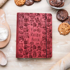May include: A red wooden recipe book with a black elastic closure. The cover is decorated with a pattern of kitchen utensils and the words "Family Recipe Book".