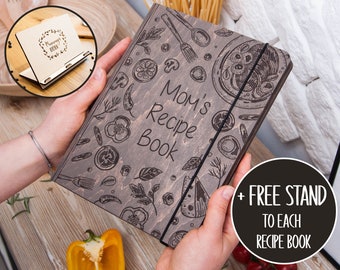 May include: A brown wooden recipe book with a black elastic band closure. The cover features a design of various kitchen utensils and ingredients, including tomatoes, peppers, and herbs. The text "Mom's Recipe Book" is engraved on the cover. A free stand is included with the book.