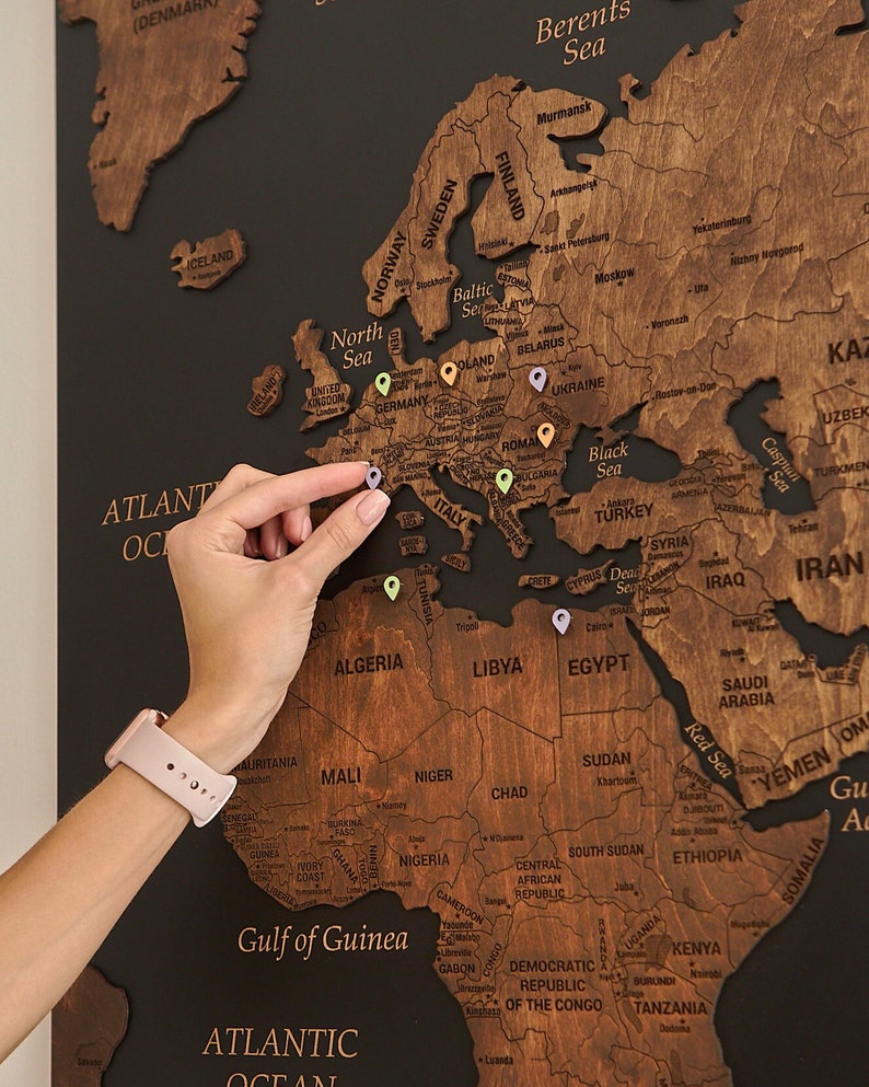 May include: A wooden world map with push pins marking locations. The map is mounted on a black background. The map is made of wood and has a dark brown stain. The push pins are various colors, including red, green, blue, and yellow.