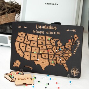 May include: A black wooden map of the United States with a corkboard surface. The map is decorated with a compass and an arrow. The text "Our adventures The Garrisons est. June 10, 2016" is printed on the map. The map is surrounded by colorful push pins.