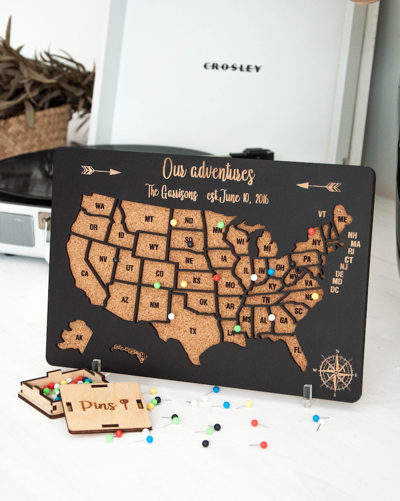 May include: A black wooden map of the United States with a corkboard surface. The map is decorated with a compass and an arrow. The text "Our adventures The Garrisons est. June 10, 2016" is printed on the map. The map is surrounded by colorful push pins.