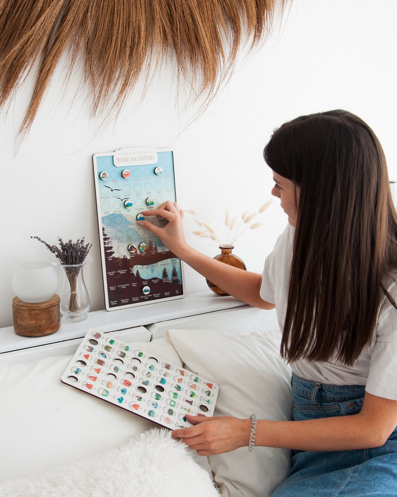 May include: A wooden board with a map of the United States National Parks. Each park is represented by a small, colorful icon. A person is holding a separate board with the same icons, but they are larger and can be used to mark which parks they have visited.