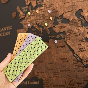 May include: Three wooden push pins in different colors: green, purple, and orange. Each pin has a grid pattern of small circles. The pins are held in a hand against a wooden world map.