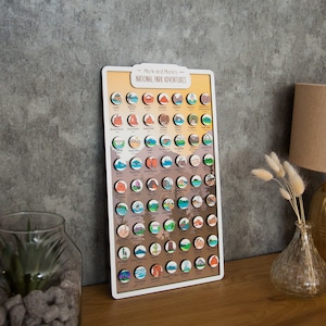 May include: A wooden board with a light brown background and white border. The board features a colorful map of the United States with circular icons representing each national park. The title "Mark and Manon's National Park Adventures" is printed at the top of the board.