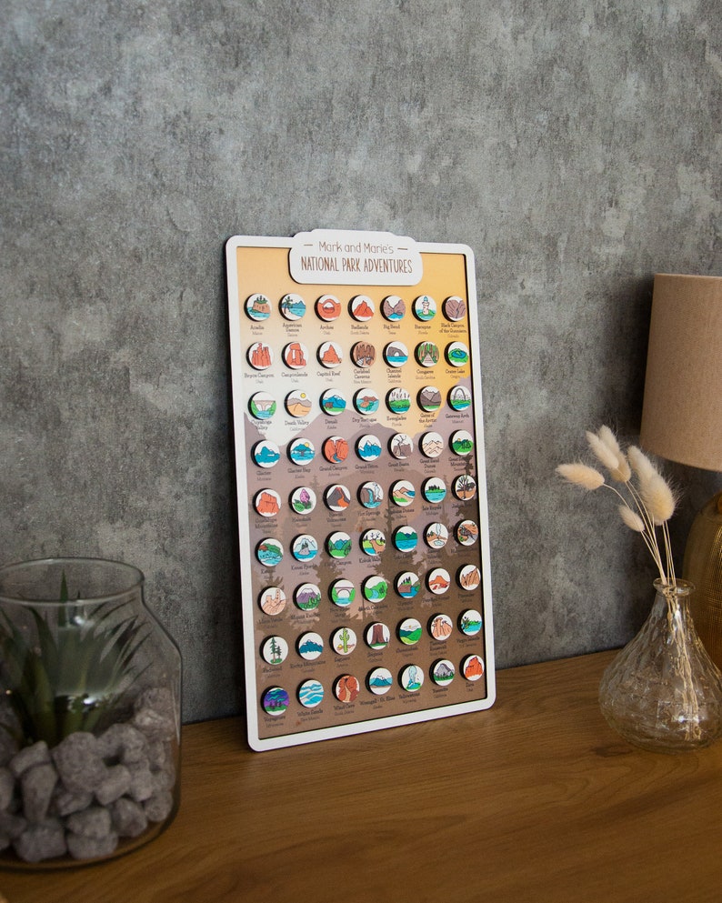May include: A wooden board with a light brown background and white border. The board features a colorful map of the United States with circular icons representing each national park. The title "Mark and Manon's National Park Adventures" is printed at the top of the board.