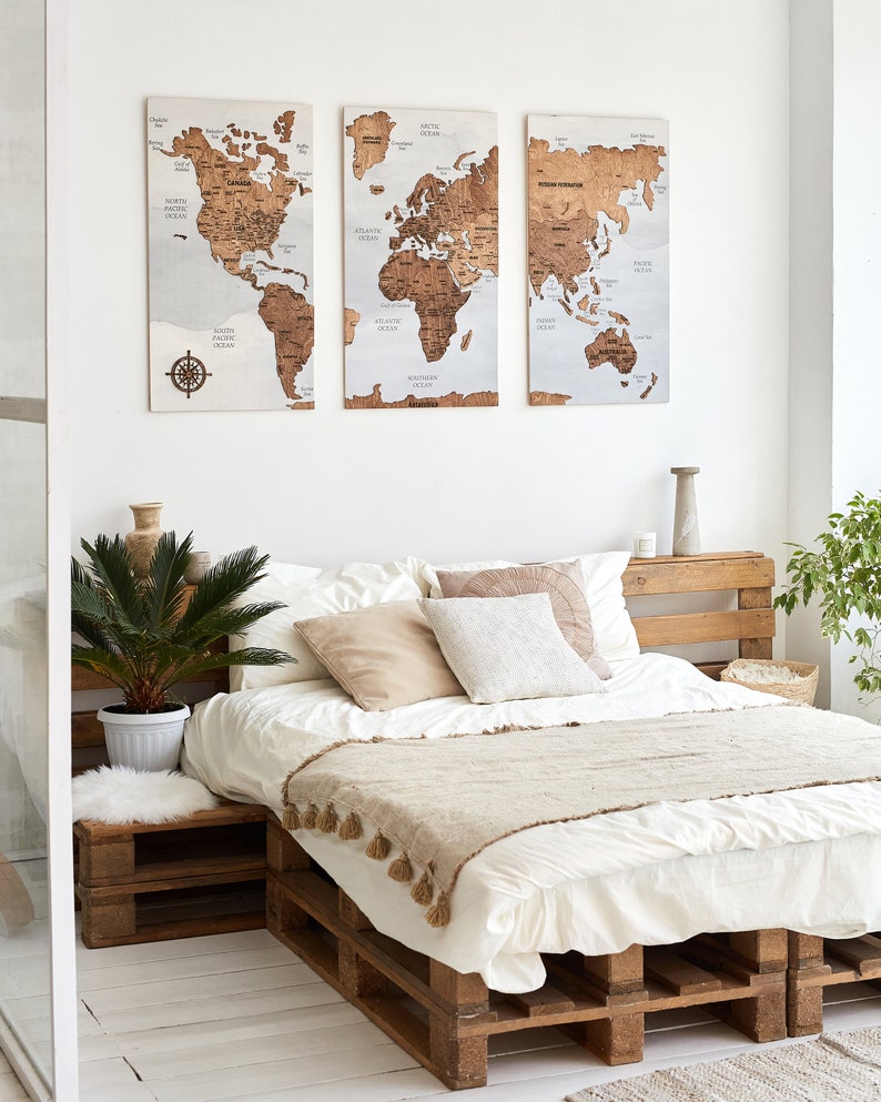 May include: A white bed frame made of wooden pallets with a white duvet and a beige throw blanket. The bed is decorated with beige and white pillows. Three wooden world maps are hanging on the wall behind the bed.