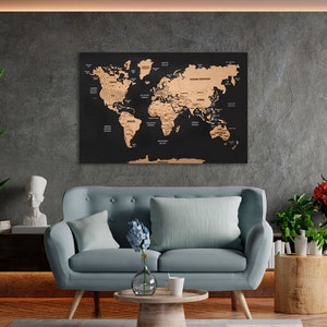 May include: A black corkboard world map with gold foil accents. The map is hanging on a gray wall in a living room setting. A light blue sofa with two pillows is in the foreground. A vase with blue flowers and a white mug are on a wooden coffee table.