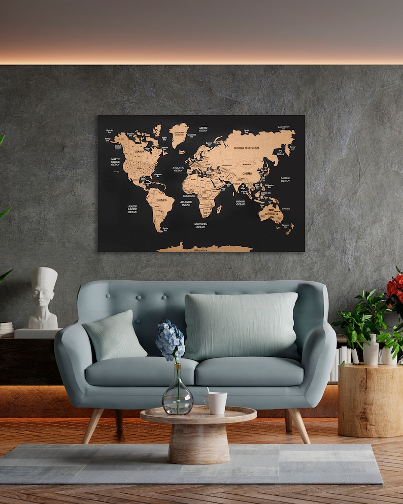 May include: A black corkboard world map with gold foil accents. The map is hanging on a gray wall in a living room setting. A light blue sofa with two pillows is in the foreground. A vase with blue flowers and a white mug are on a wooden coffee table.