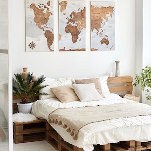 May include: A white bed with a wooden pallet frame and a beige throw blanket. The bed is decorated with beige and white pillows. Three wooden world maps are hanging on the wall behind the bed.