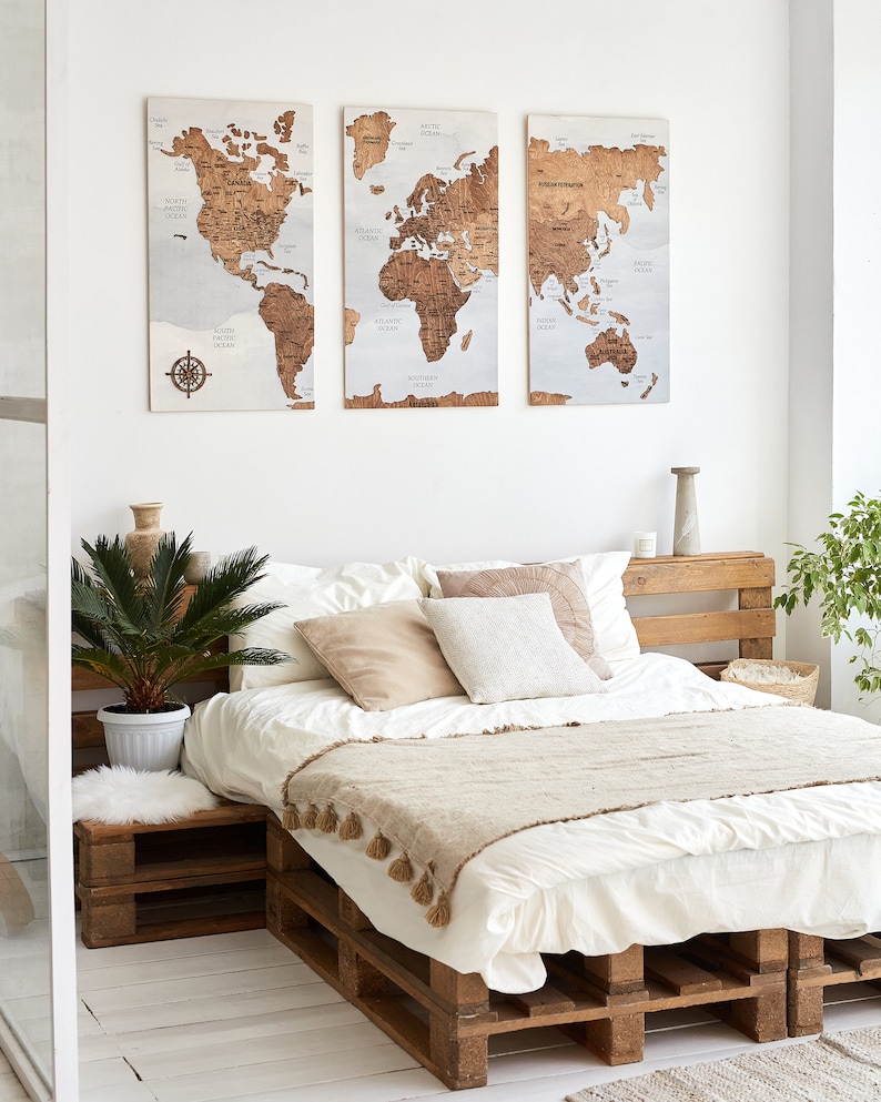 May include: A white bed with a wooden pallet frame and a beige throw blanket. The bed is decorated with beige and white pillows. Three wooden world maps are hanging on the wall behind the bed.