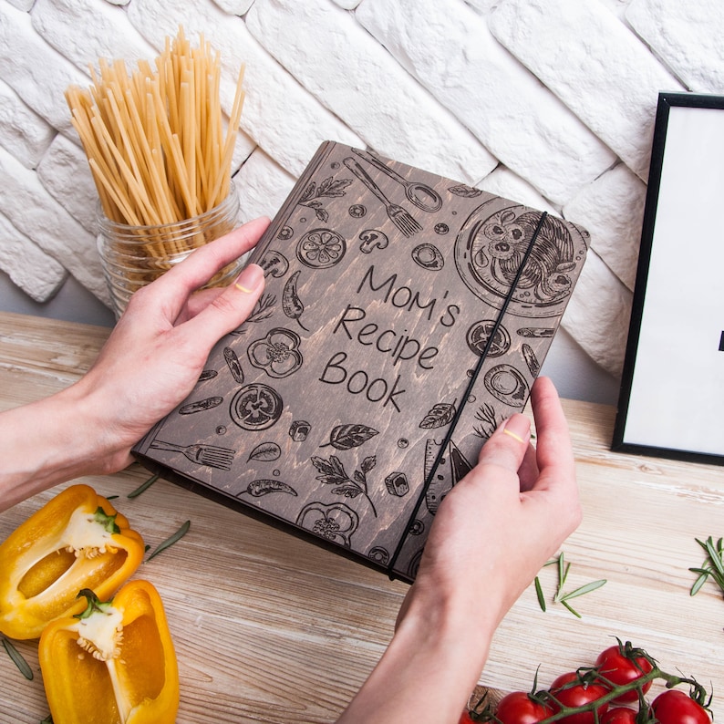 May include: A brown wooden recipe book with a black elastic closure. The cover is decorated with a black and white illustration of kitchen utensils, food, and the text "Mom's Recipe Book".