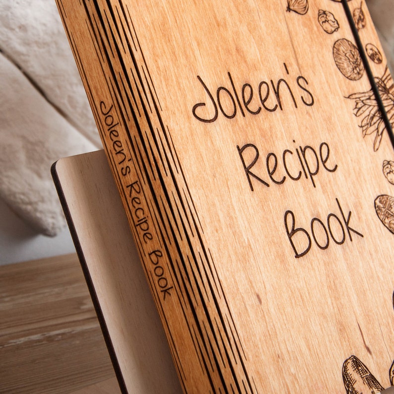 May include: A wooden recipe book with the name "Joleen's Recipe Book" engraved on the cover. The book has a rustic look and is perfect for storing favorite recipes.