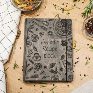 May include: A gray wooden recipe book with a black elastic band closure. The cover features a hand-drawn design of kitchen utensils, vegetables, and pasta. The text "Janet's Recipe Book" is engraved on the front.