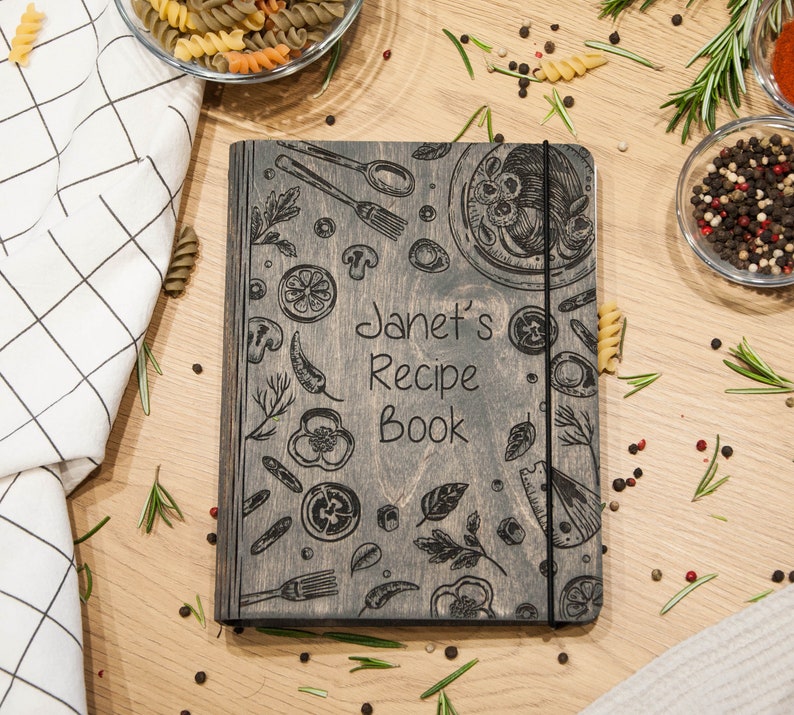 May include: A gray wooden recipe book with a black elastic band closure. The cover features a hand-drawn design of kitchen utensils, vegetables, and pasta. The text "Janet's Recipe Book" is engraved on the front.