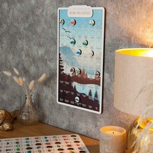 May include: A white board with the text "Mark and Marie's National Park Adventures" and a map of the United States with circles for each national park. The board has a white frame and is hanging on a gray wall. There are small, colorful stickers on the board, representing each national park.