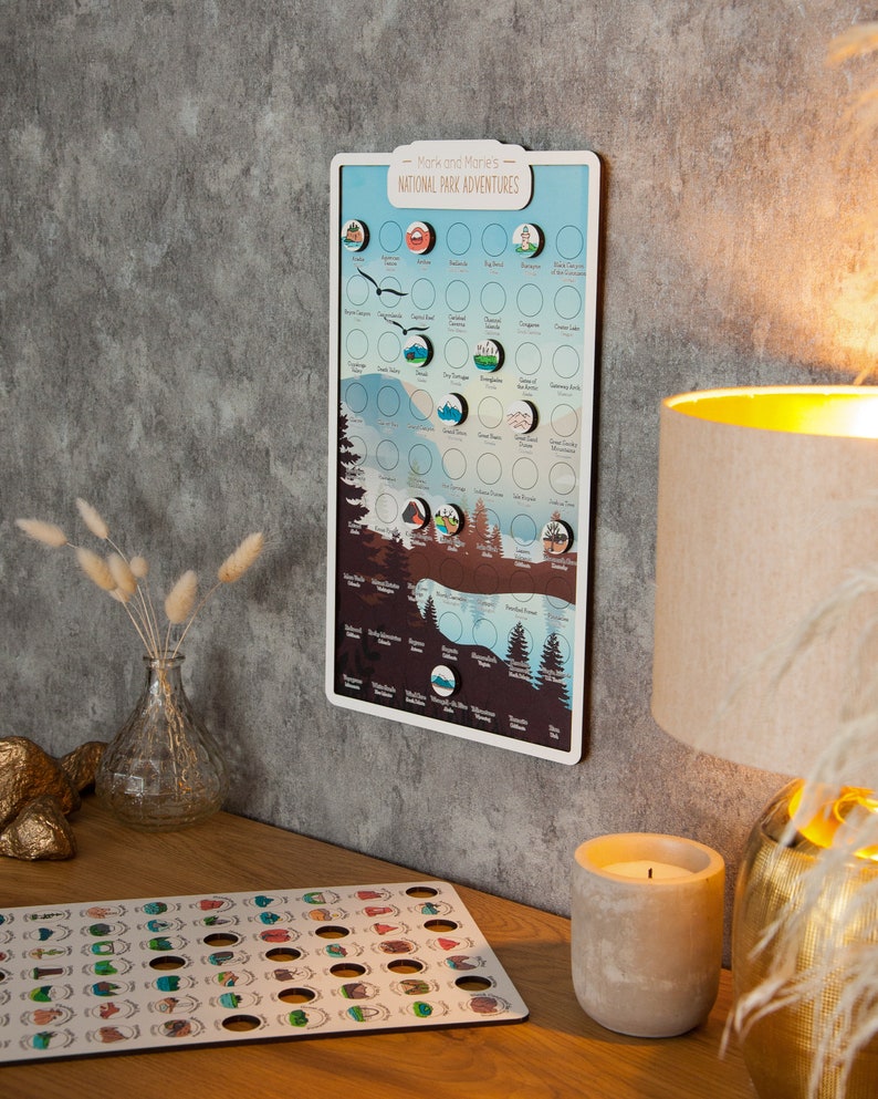 May include: A white board with the text "Mark and Marie's National Park Adventures" and a map of the United States with circles for each national park. The board has a white frame and is hanging on a gray wall. There are small, colorful stickers on the board, representing each national park.