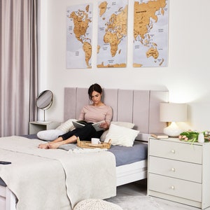 May include: A woman sits on a bed with a white blanket and gray pillows, reading a book. A white dresser with three drawers is on the right side of the image. Three world maps are hanging on the wall behind the bed. The maps are made of cork and have a light brown color.
