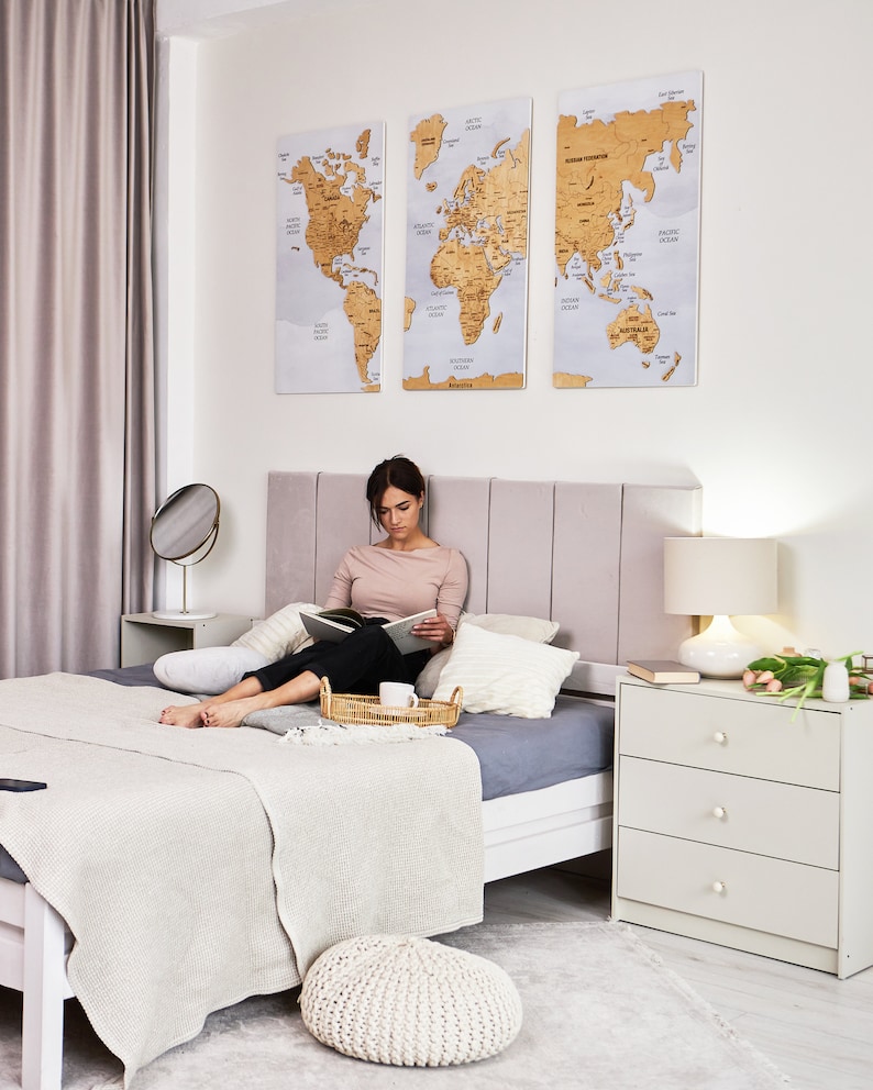 May include: A woman sits on a bed with a white blanket and gray pillows, reading a book. A white dresser with three drawers is on the right side of the image. Three world maps are hanging on the wall behind the bed. The maps are made of cork and have a light brown color.