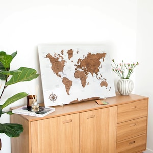 Puede incluir: Un mapa del mundo de madera con continentes marrones sobre un fondo blanco. Una rosa de los vientos está presente. El mapa se muestra en un mueble de madera clara, junto a un jarrón con flores y una planta.
