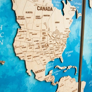 May include: A wooden map of North America with the states and provinces labeled. The map is on a blue background with a watercolor effect. The map is divided into two sections, with the eastern section showing the Atlantic Ocean and the western section showing the Pacific Ocean.
