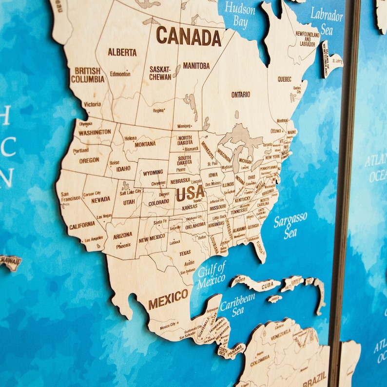 May include: A wooden map of North America with the states and provinces labeled. The map is on a blue background with a watercolor effect. The map is divided into two sections, with the eastern section showing the Atlantic Ocean and the western section showing the Pacific Ocean.