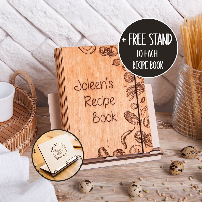 May include: A personalized wooden recipe book with the name "Joleen's Recipe Book" engraved on the cover. The book features a stand and is decorated with a wood-burned design of various vegetables and herbs. The text "+ FREE STAND TO EACH RECIPE BOOK" is displayed in a black circle.
