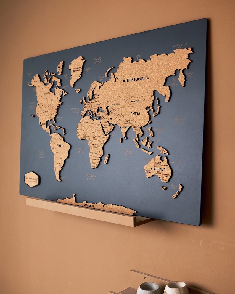 May include: A cork world map with a blue background. The map is mounted on a wooden shelf. The map is made of cork and has a raised design. The map is labeled with the names of continents and oceans.
