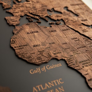 May include: A close-up of a wooden world map with the countries of Africa highlighted. The map is mounted on a dark gray background. The text "Gulf of Guinea" and "Atlantic Ocean" are visible.