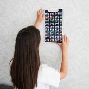 May include: A person is hanging a poster on a wall. The poster is a National Park Adventures scratch-off map. The map is black with white and colored circles that represent different national parks. The map has the text "National Park Adventures" at the top.