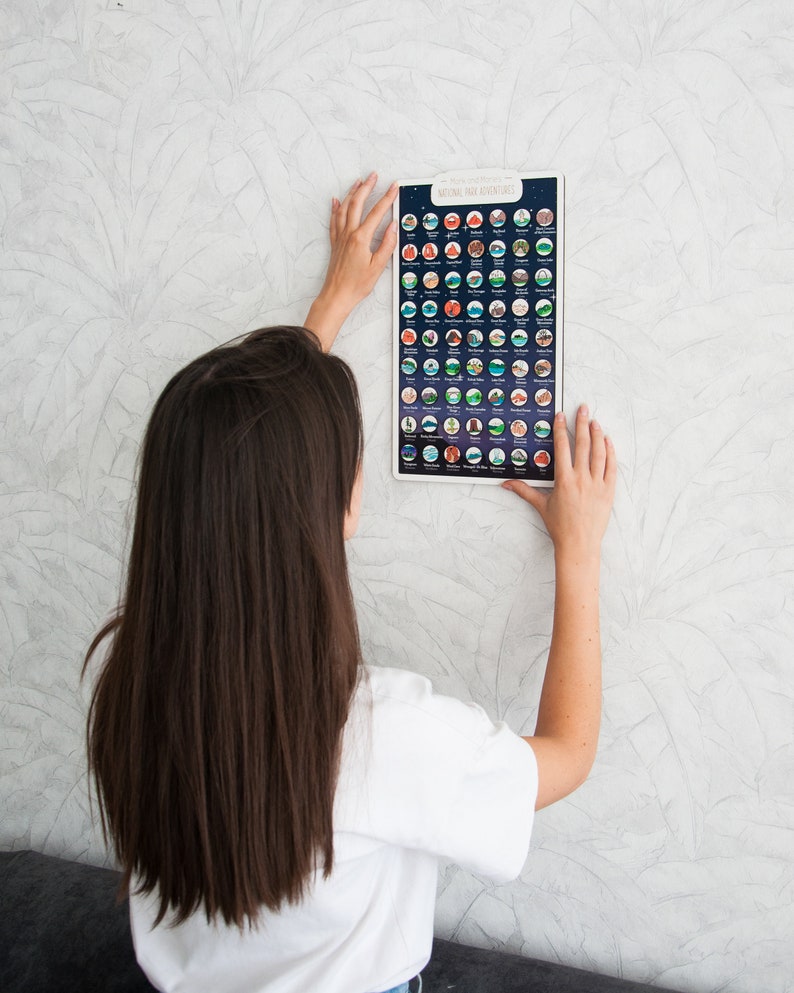 May include: A person is hanging a poster on a wall. The poster is a National Park Adventures scratch-off map. The map is black with white and colored circles that represent different national parks. The map has the text "National Park Adventures" at the top.
