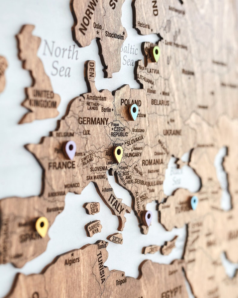 May include: A wooden world map with push pins marking various locations in Europe. The map is made of light brown wood and has a white background. The map includes countries such as Norway, Sweden, Finland, Denmark, Germany, Poland, Czech Republic, Slovakia, Austria, Hungary, Romania, Italy, France, Spain, Portugal, and more.