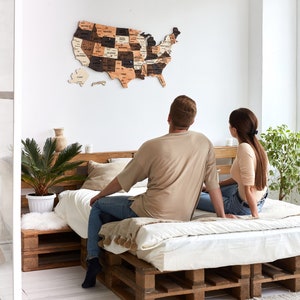 May include: A couple sits on a bed made from wooden pallets, looking at a wooden map of the United States hanging on the wall. The map is made up of individual state pieces, each with a different wood grain and colour. The couple is looking at the map, possibly planning a trip.
