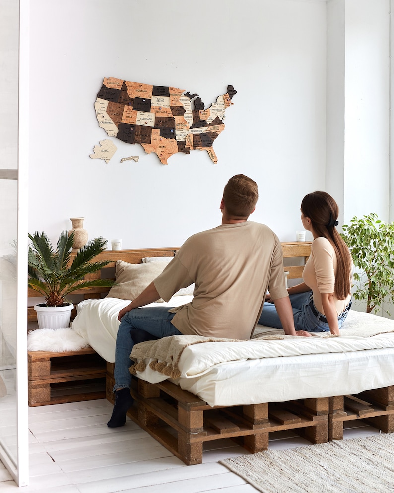 May include: A couple sits on a bed made from wooden pallets, looking at a wooden map of the United States hanging on the wall. The map is made up of individual state pieces, each with a different wood grain and colour. The couple is looking at the map, possibly planning a trip.