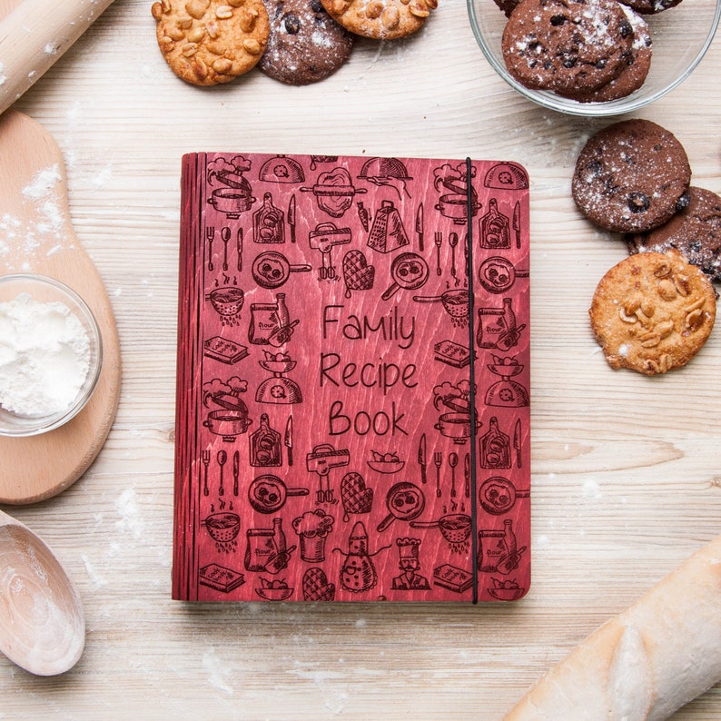 May include: A red wooden recipe book with a black elastic closure. The cover is decorated with a pattern of kitchen utensils and the words "Family Recipe Book".