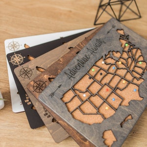 May include: A set of four wooden US map push pin boards with cork surfaces. The boards are in different shades of brown and gray. The top board has the text "Adventure Awaits" engraved on it. The boards are stacked on top of each other.