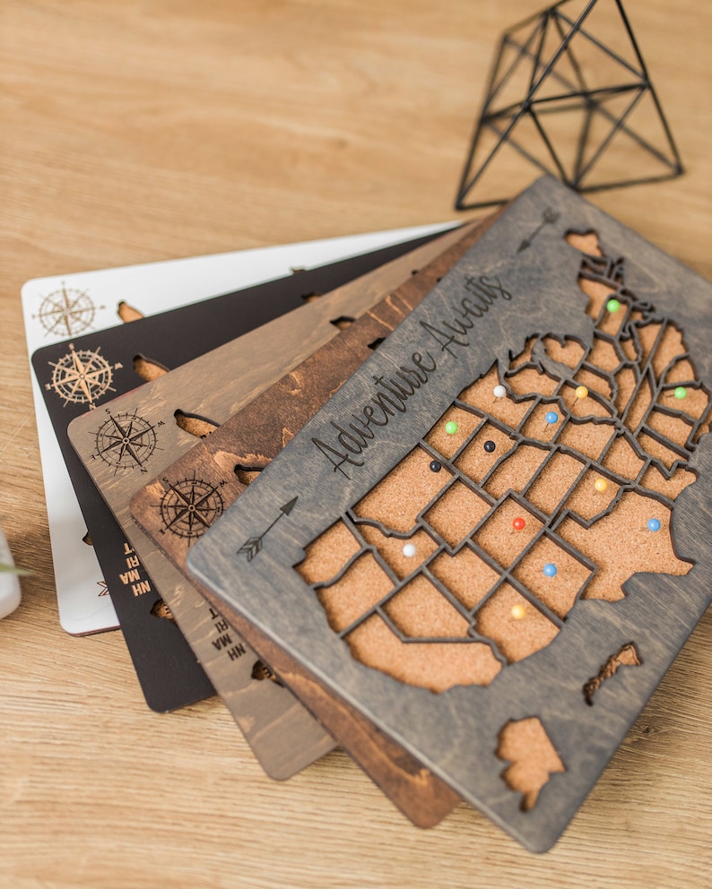 May include: A set of four wooden US map push pin boards with cork surfaces. The boards are in different shades of brown and gray. The top board has the text "Adventure Awaits" engraved on it. The boards are stacked on top of each other.