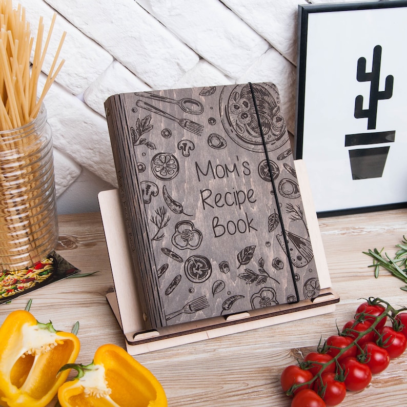 May include: A gray wooden recipe book with a black elastic band closure. The cover features a hand-drawn design of various food items, including vegetables, herbs, and a pizza. The text "Mom's Recipe Book" is engraved on the cover.