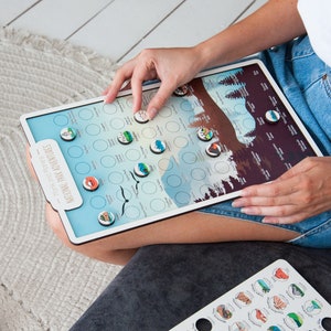 May include: A white board with a colorful map of National Parks in the United States. The map is divided into circles with icons representing each park. A person is placing a small wooden icon on the map.