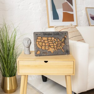 May include: A wooden map of the United States with corkboard surface and push pins. The map is mounted on a stand and has the text "Adventure Awaits" at the top. The map is sitting on a wooden end table with a drawer.