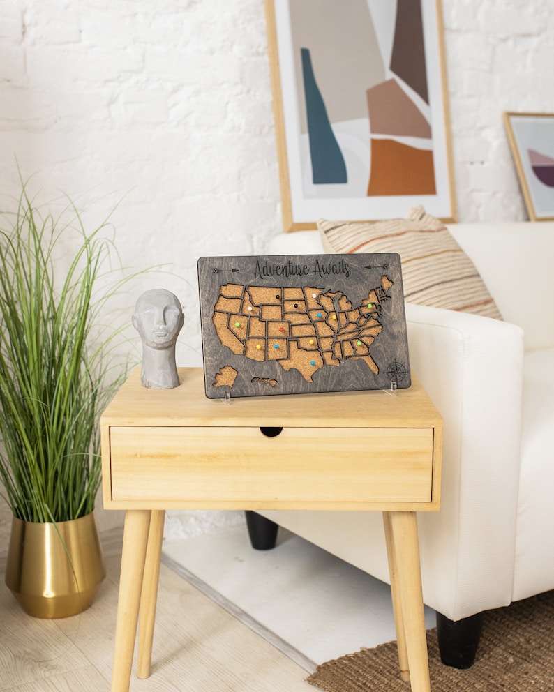 May include: A wooden map of the United States with corkboard surface and push pins. The map is mounted on a stand and has the text "Adventure Awaits" at the top. The map is sitting on a wooden end table with a drawer.