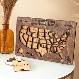 May include: A wooden map of the United States with corkboard surface and push pins. The map is on a stand and has the text "An Unexpected Journey Appgood Family est. April 27, 2019". A small wooden box with the text "Pins &" is in the foreground.