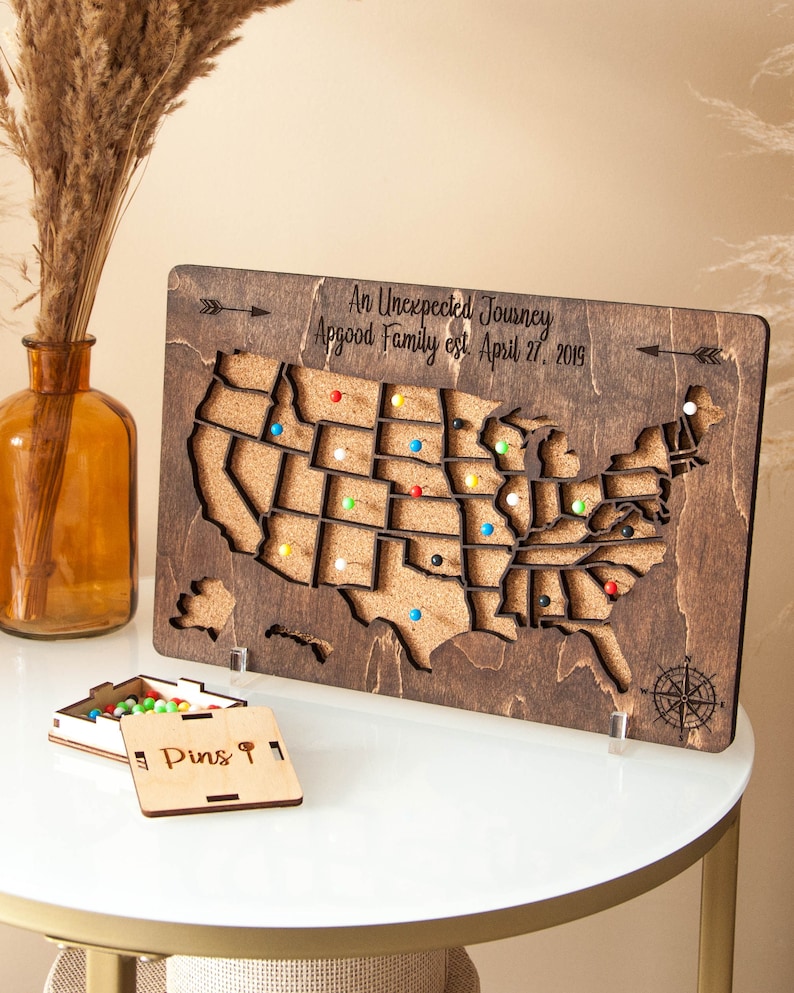 May include: A wooden map of the United States with corkboard surface and push pins. The map is on a stand and has the text "An Unexpected Journey Appgood Family est. April 27, 2019". A small wooden box with the text "Pins &" is in the foreground.